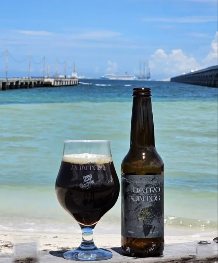 Cuatro Puntos Oscura servida en copa junto a la botella frente al muelle